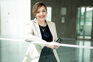 Prof. Asta Pundzėnienė šypsosi šviesioje, modernioje aplinkoje, apsirengusi šviesiu švarku, rankoje laikydama telefoną.