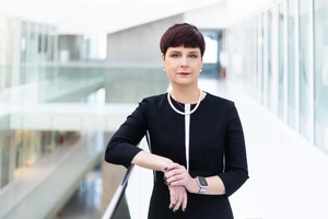 Prof. Edita Gimžauskienė stovi moderniame stiklo koridoriuje, vilkėdama juodą suknelę su baltu apvadu.